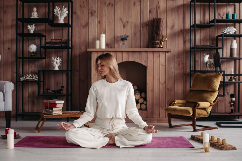 A person in white clothing sits cross-legged on a purple mat in a cozy room with wooden walls and a serene ambiance.