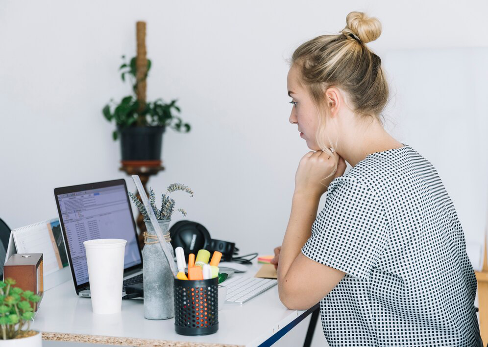 A person in a black and white patterned shirt works at a desk with a laptop, stationery,
