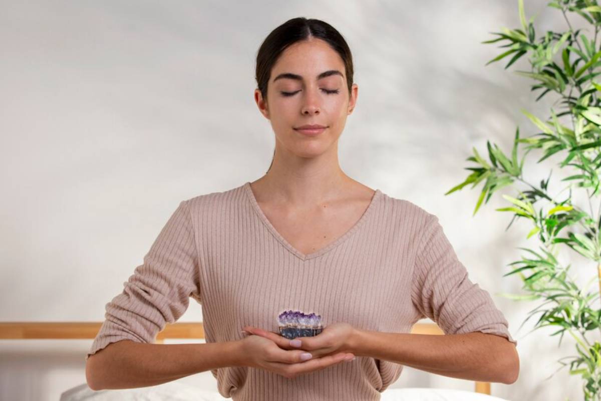 A person in a beige sweater holds a small crystal in their hands, surrounded by soft light and greenery in a serene indoor setting.