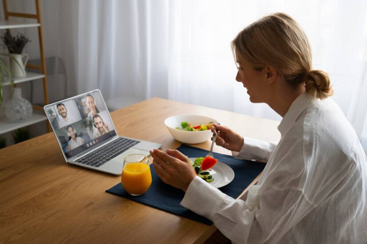 A person enjoys a meal at a wooden table while participating in a video call on their laptop, with a bowl of salad nearby.