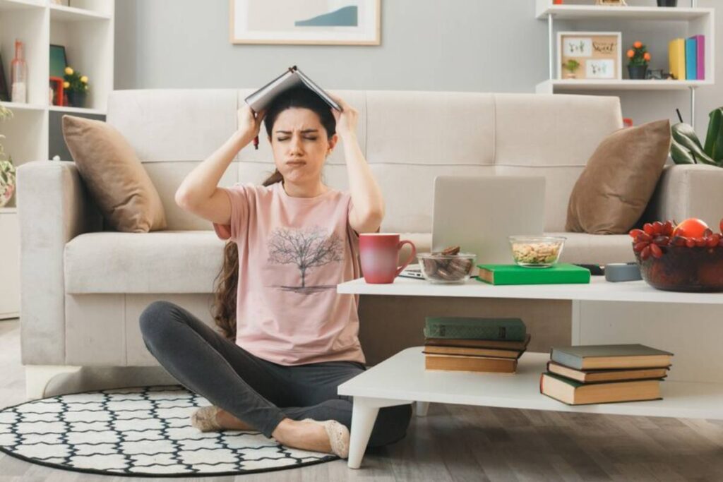 A person sits on the floor of a cozy living room looking stressed, holding a book over their head, surrounded by snacks and a laptop.