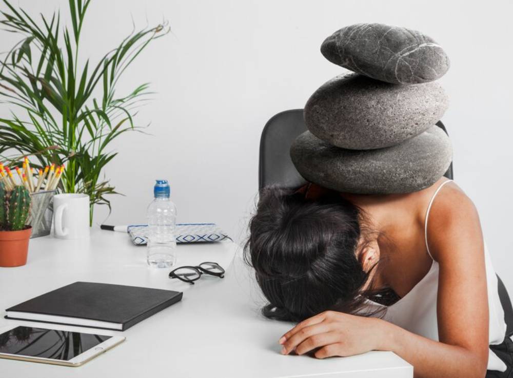 A stressed woman rests her head on a desk, weighted down by stacked stones, in a cluttered office environment.
