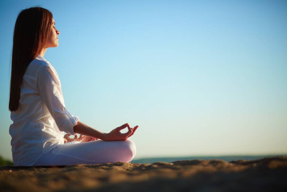 A person sits in a meditative pose on a sandy surface, with a clear blue sky in the background, promoting a sense of tranquility.