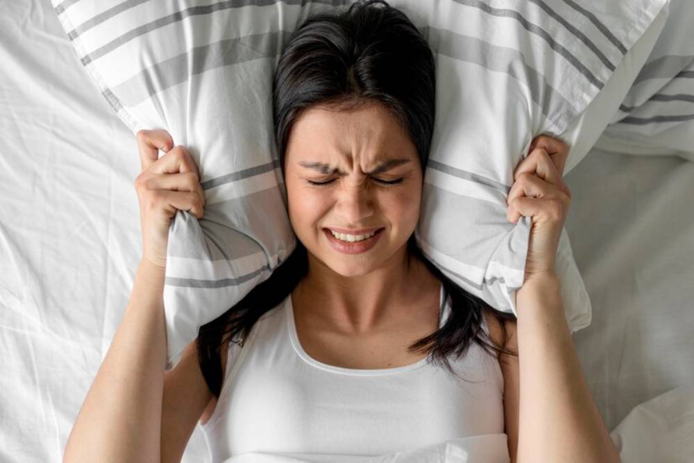 A stressed person lies in bed, grasping two pillows tightly against their head, surrounded by white bedding with gray stripes.