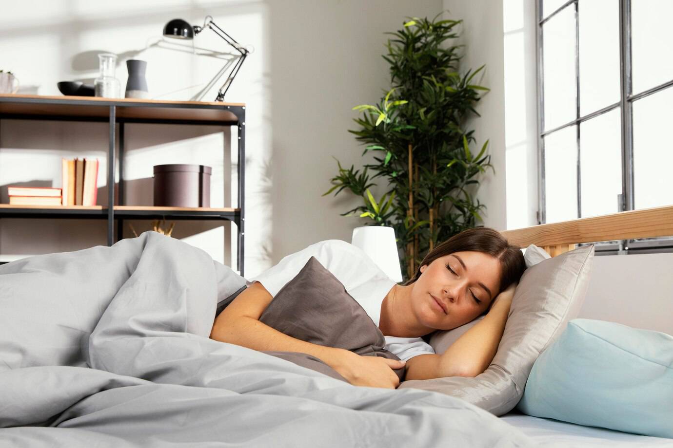 A person rests peacefully in bed, surrounded by soft gray blankets and pillows, with a modern room featuring greenery and natural light.